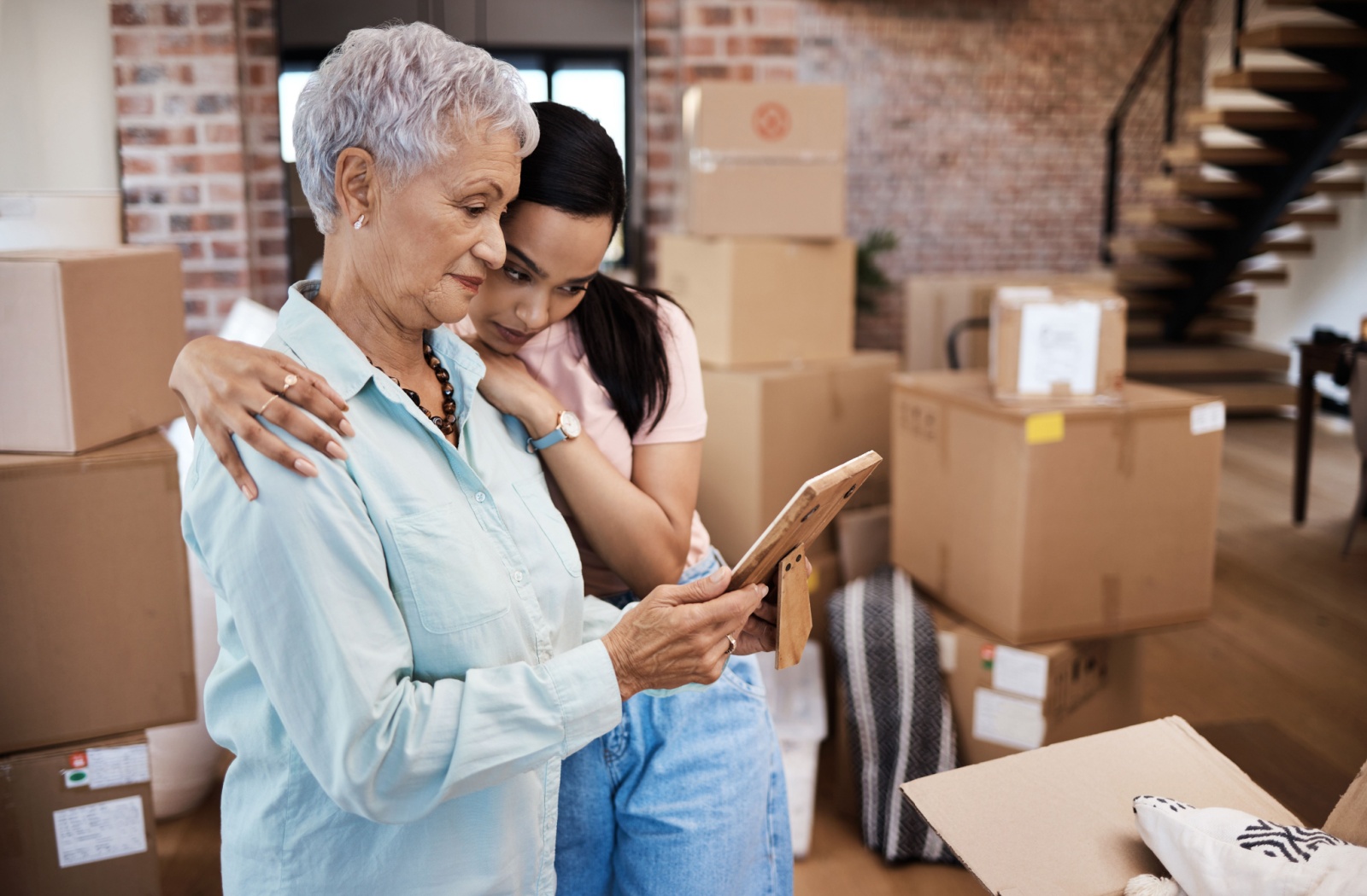 A senior and their adult child hugging from the side and looking at a framed photo while surrounded by moving boxes.
