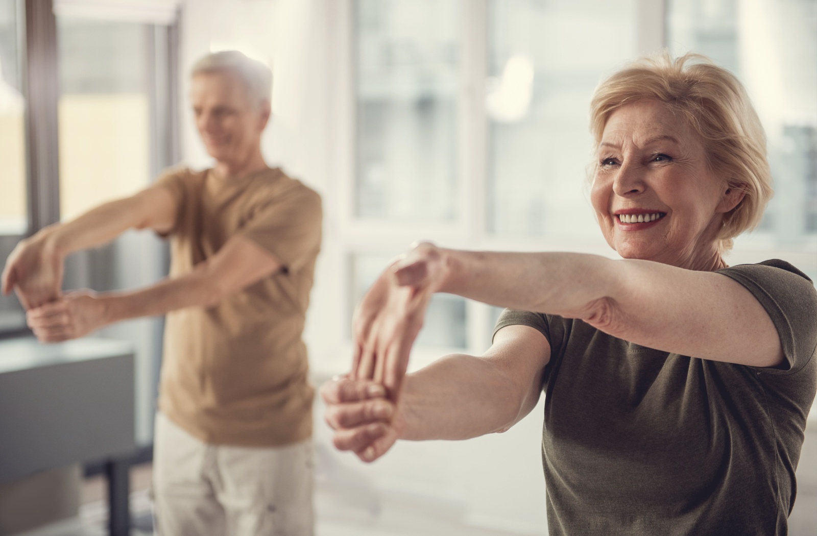 2 older adults smile while stretching their hands downward to improve their flexibility