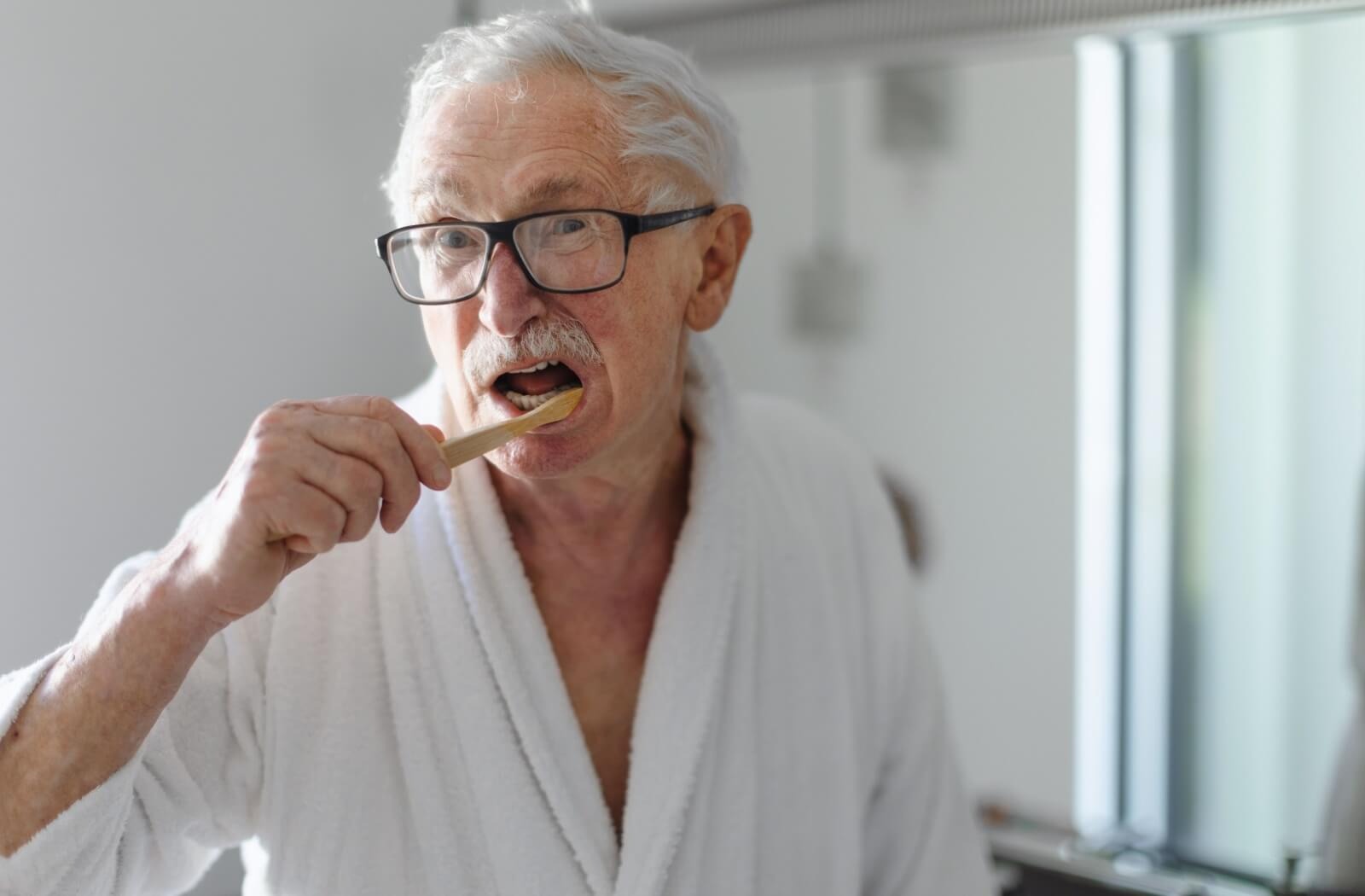 An older adult in a white robe brushes their teeth in the morning
