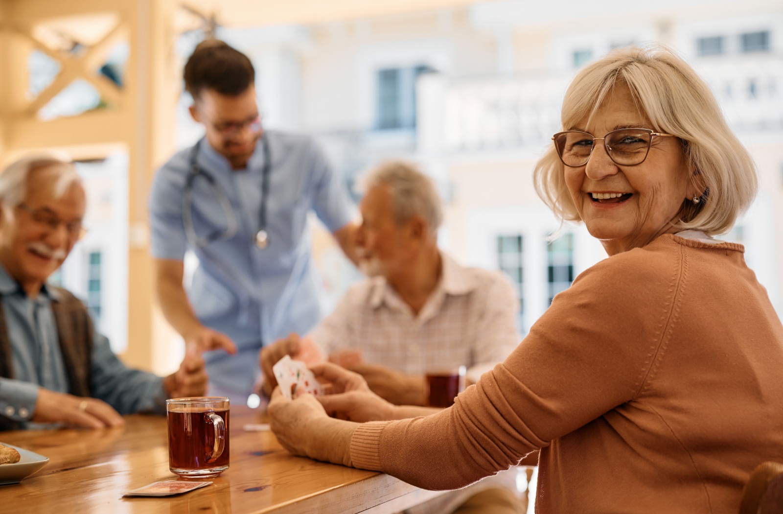 A senior woman sits at a table, holding cards next to a cup of tea and smiling. In the background, fellow seniors engage in conversation while a nurse helps out.
