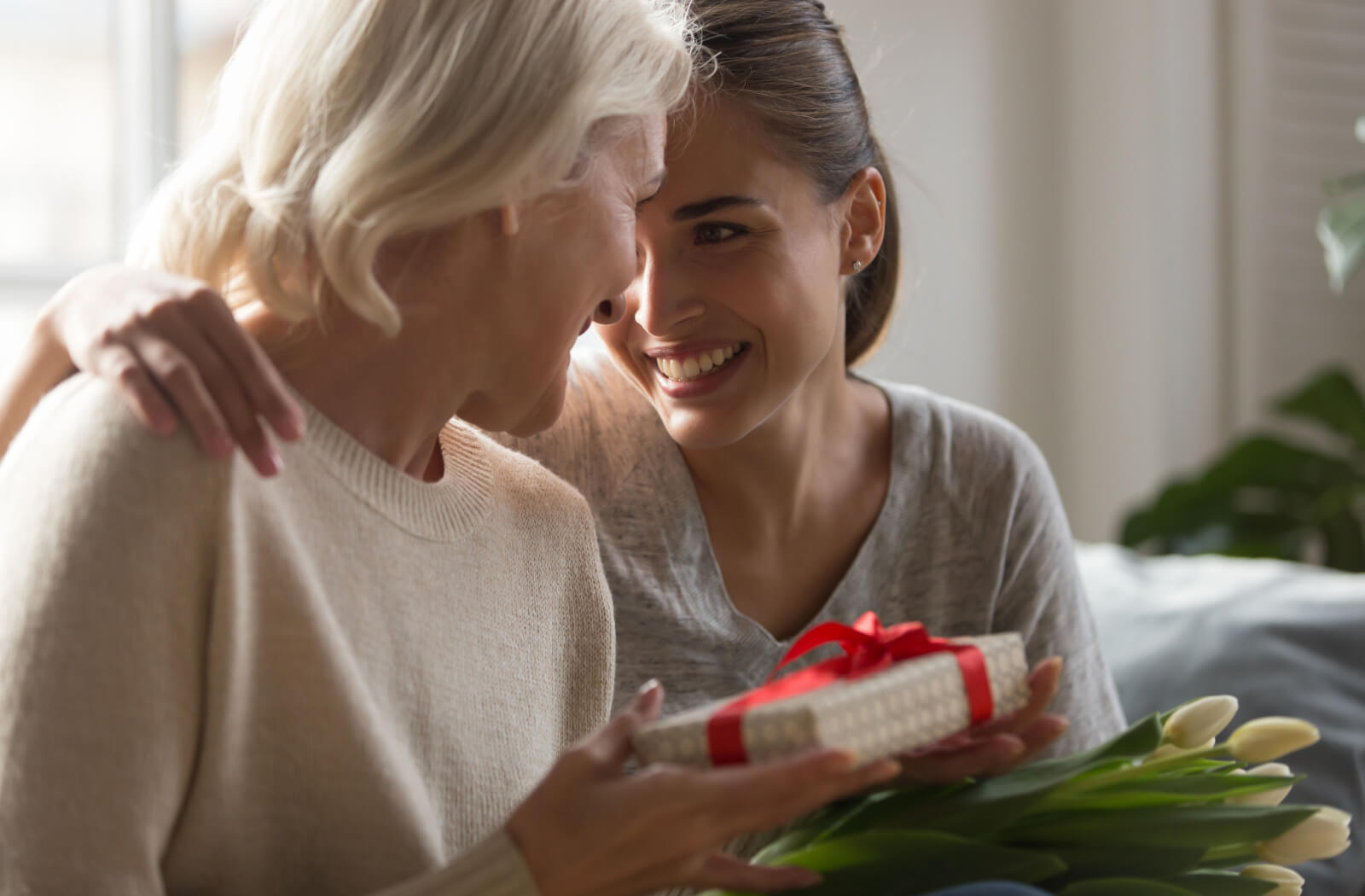 An adult hugging their older parent with dementia and smiling after giving a wrapped gift and flowers.