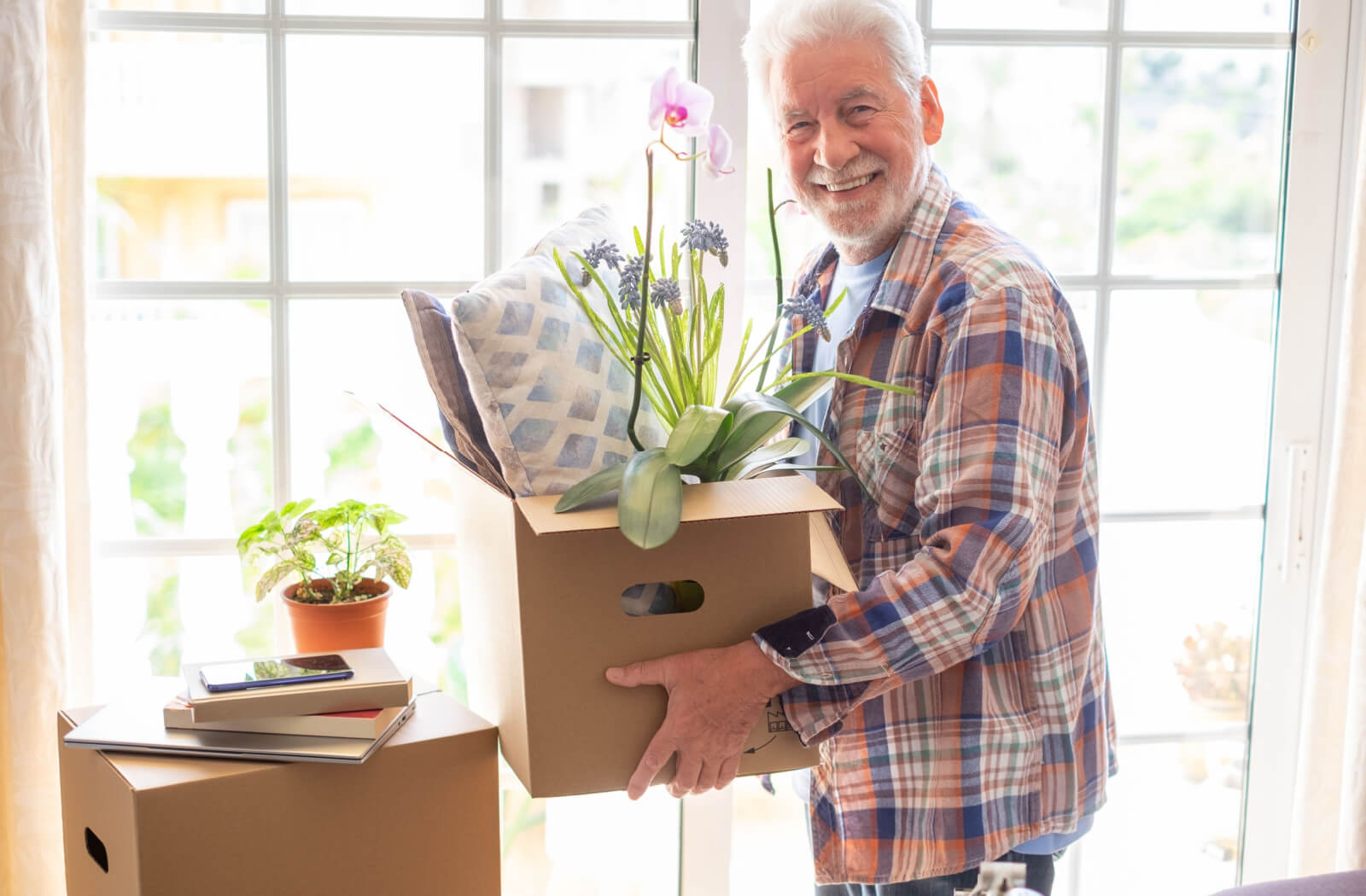 A smiling older adult holding a box of assorted household items in front of a sunlit window preparing for a move to assisted living.