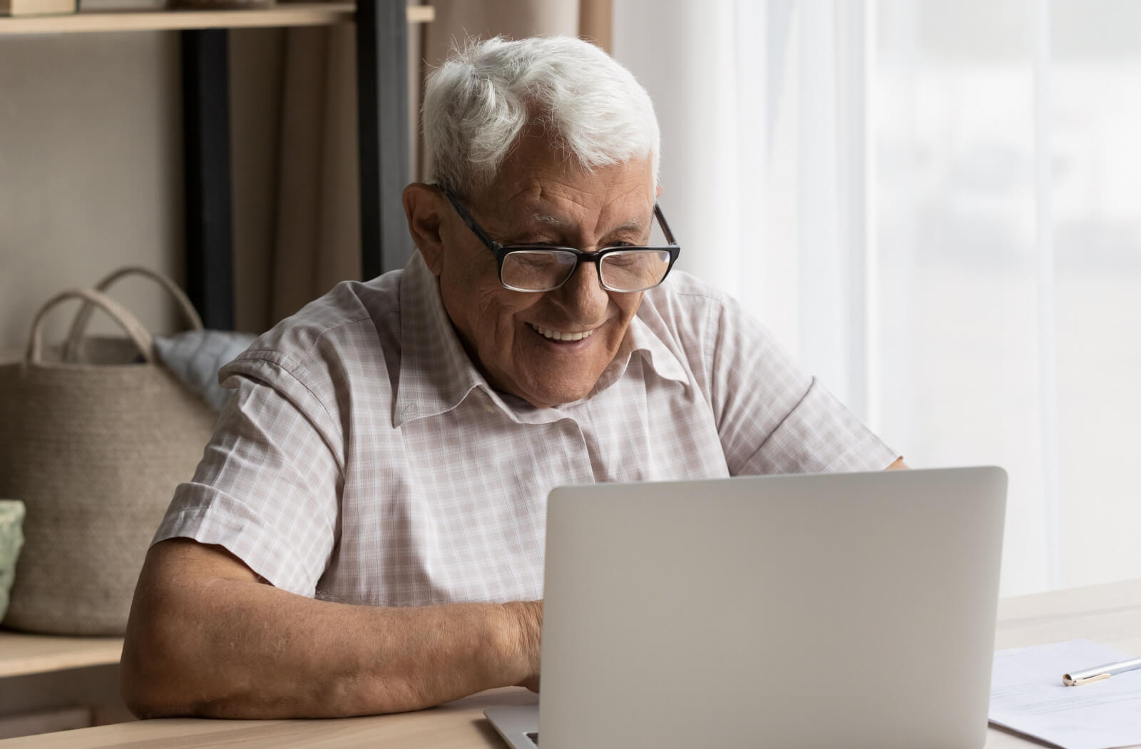 An older adult at home in front of his laptop beside a window, smiling as he's safely online.