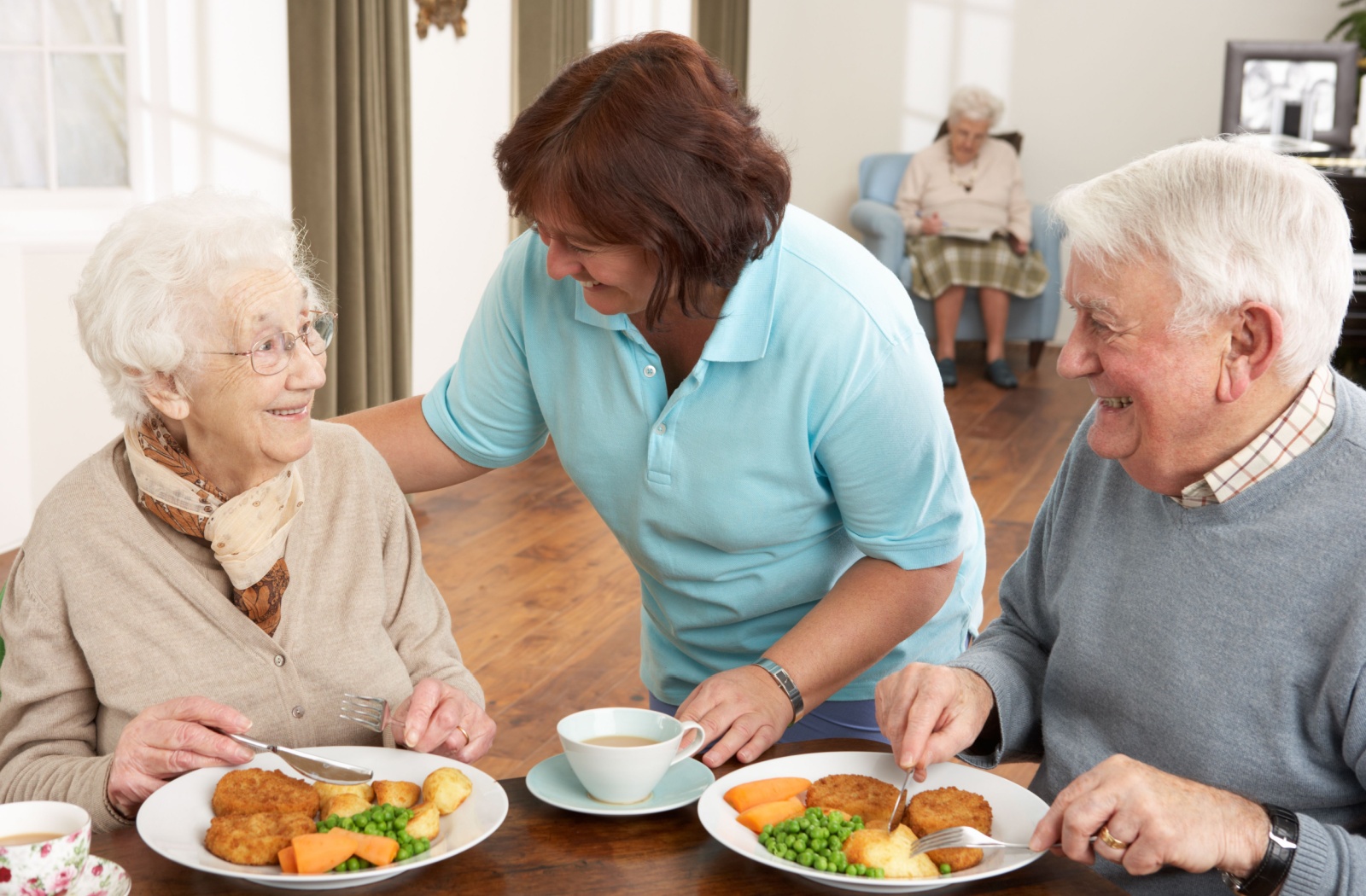 A staff member in senior living talking to a smiling older adult while they having a meal with their spouse.
